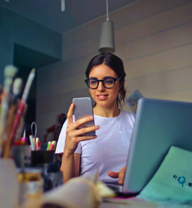 A woman with glasses looking at a cell phone while sitting in front of a laptop in an art studio.